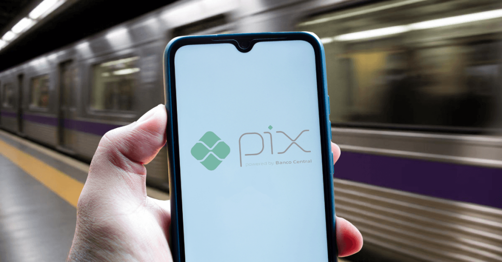 Mão segurando um smartphone com o logotipo do PIX na tela, em uma plataforma do Metrô de São Paulo com um trem em movimento desfocado ao fundo.