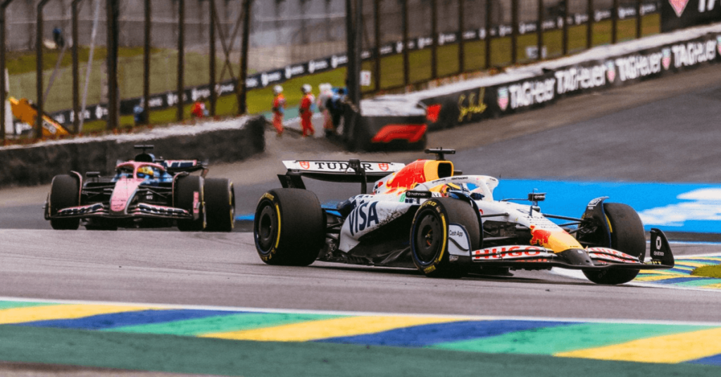 Carros de Fórmula 1 disputando posição nas clássicas zebras verde e amarelas do Autódromo de Interlagos durante o Grande Prêmio de São Paulo.
