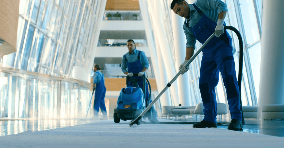 Três profissionais de limpeza com uniformes azuis operando equipamentos industriais, como aspirador e lavadora de piso, no saguão espelhado de um prédio corporativo, representando as inovações do setor na Higiexpo.